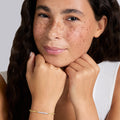A close-up lifestyle shot of a woman with freckles resting her chin on her hands, wearing a delicate gold chain bracelet with a row of diamonds on the wrist.