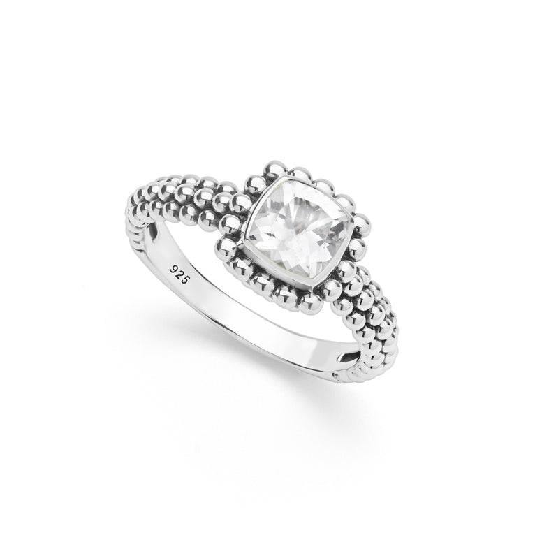 A silver ring featuring a cushion-cut white topaz gemstone surrounded by a beaded halo, with a textured beaded band, shown on a white background. 