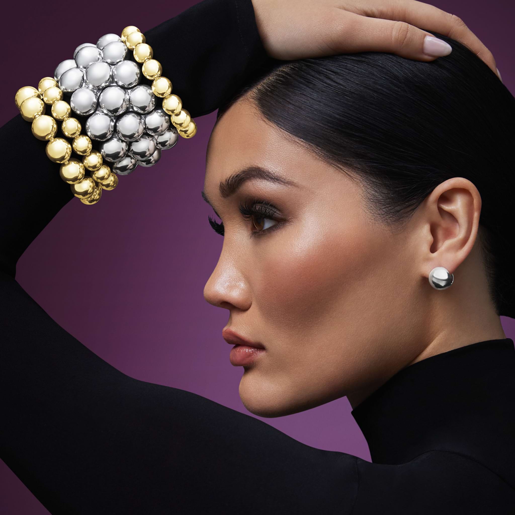 Woman wearing large silver earrings and a LAGOS Caviar Gold 18K Gold Bead Bracelet 12mm, showcasing sculptural metallic jewelry against a purplish background.