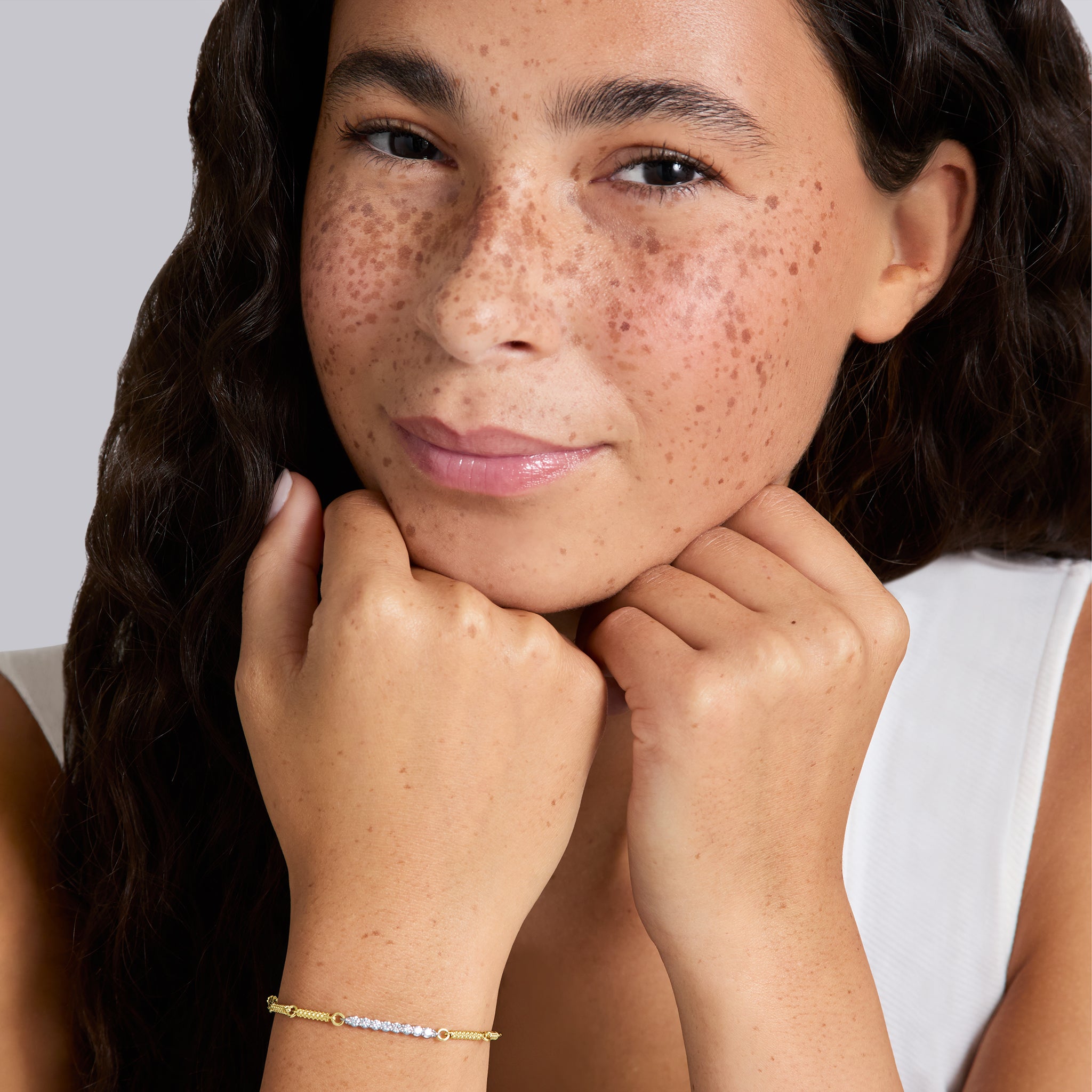 A close-up lifestyle shot of a woman with freckles resting her chin on her hands, wearing a delicate gold chain bracelet with a row of diamonds on the wrist. 