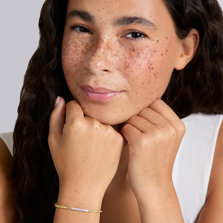 A close-up lifestyle shot of a woman with freckles resting her chin on her hands, wearing a delicate gold chain bracelet with a row of diamonds on the wrist. 