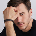 Man with hand on face wearing a black leather bracelet against a gray background