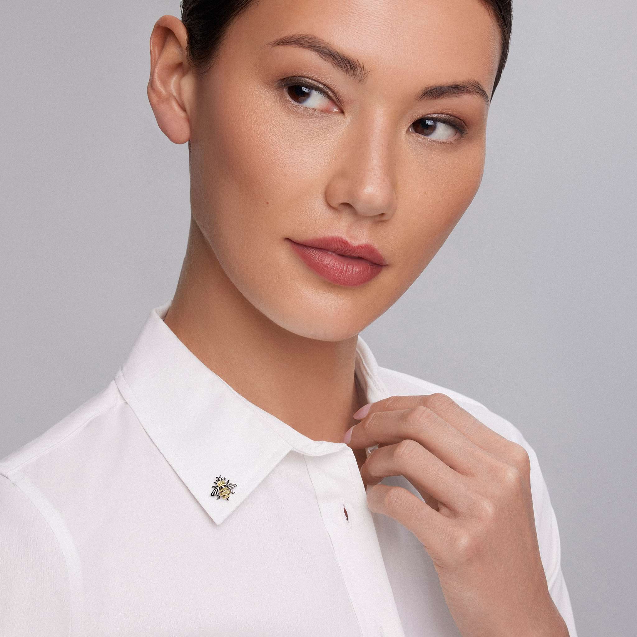 Woman wearing a crisp white shirt touches the collar adorned with the Rare Wonders Small Honeybee Pin featuring intricate 18K gold beading and black rhodium accents.
