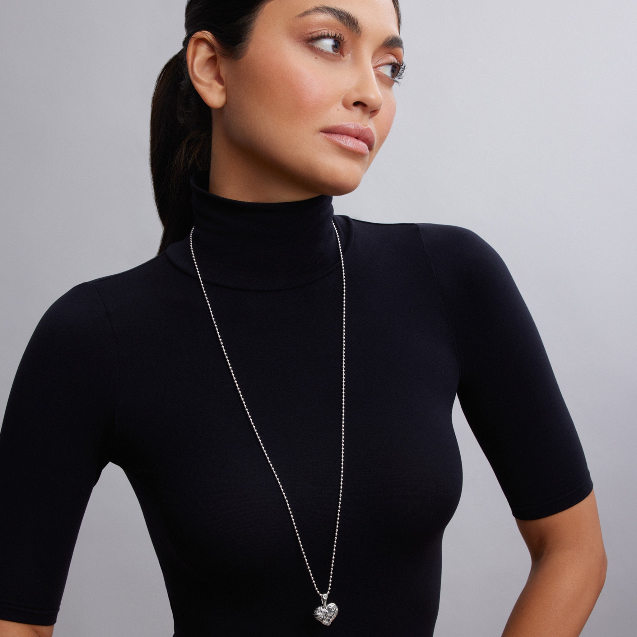 Woman wearing a black turtleneck and a long necklace with a heart pendant against a gray background