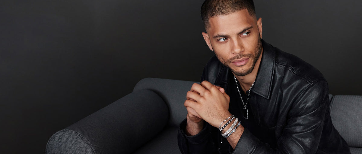 A man wearing a black leather jacket and a silver chain necklace with a dog tag, accessorized with multiple silver bracelets on one wrist, posed against a dark studio background.